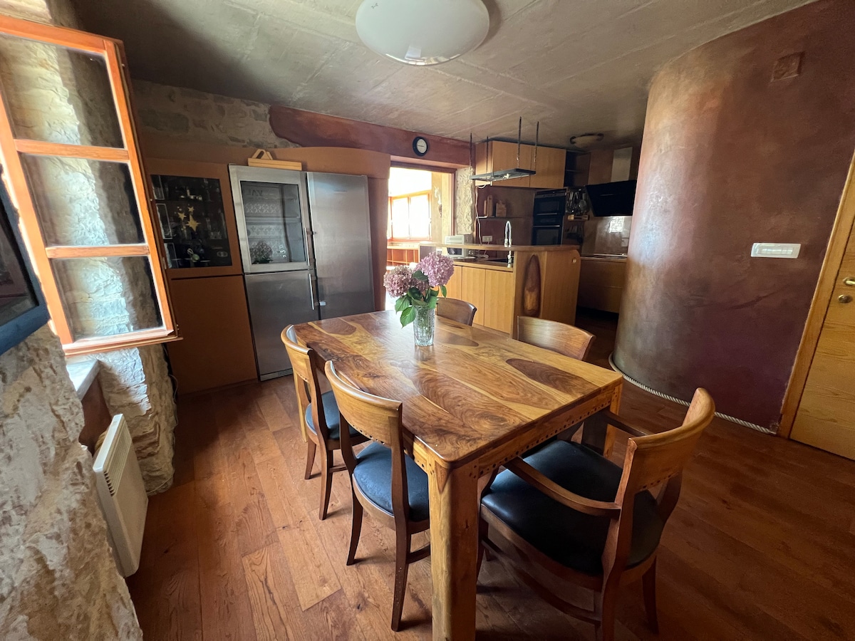 A dining area is featured with a wooden table surrounded by six chairs. Natural light enters through a window with orange frames. The kitchen area is visible in the background, showcasing modern appliances and a blend of rustic elements.