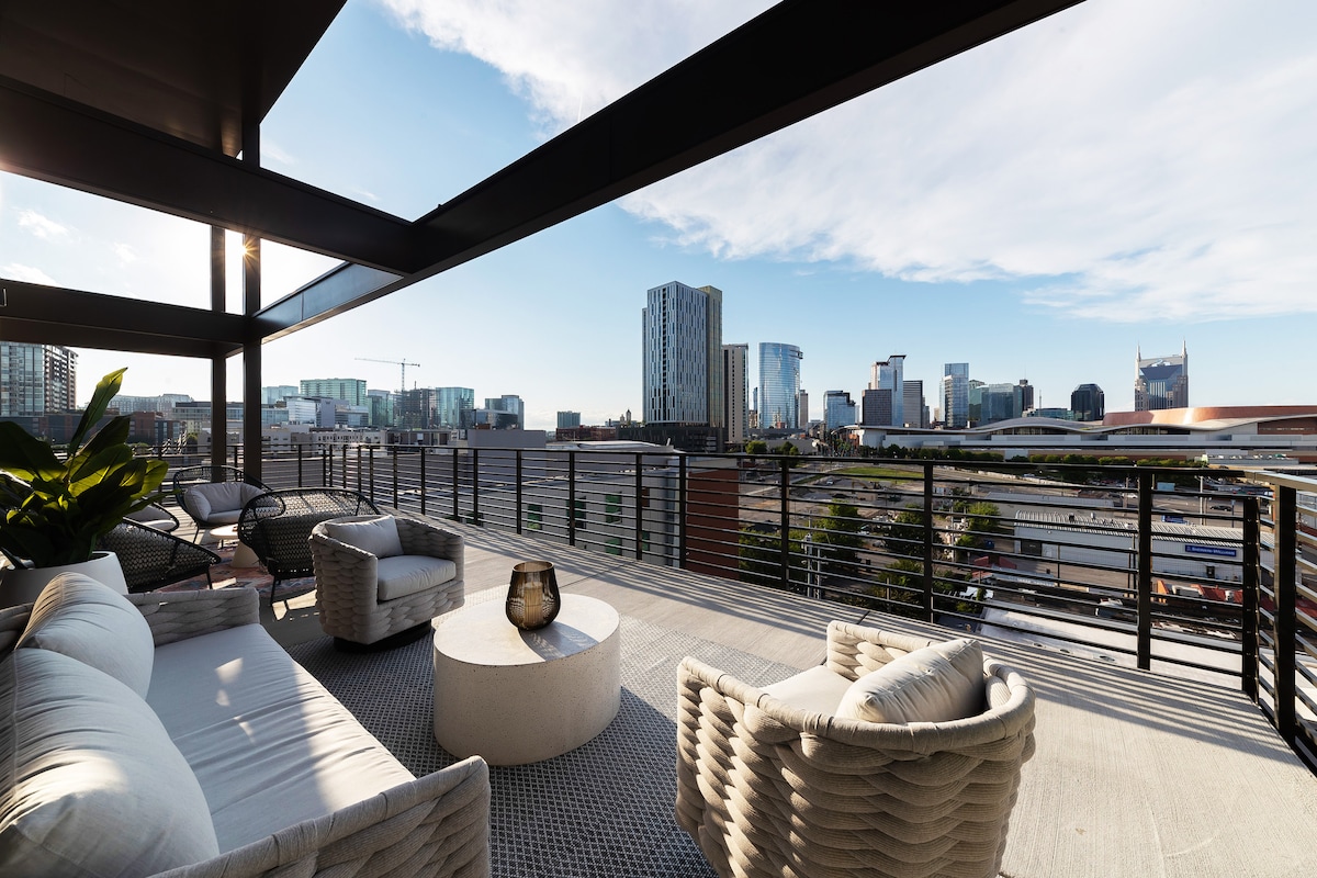 A private balcony space is showcased with modern lounge furniture, featuring a circular table and a comfortable sofa. The skyline of Nashville is visible in the background, illuminated by natural light, highlighting the urban landscape and architectural details.