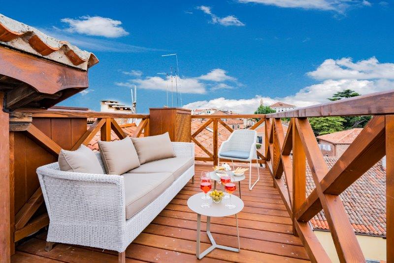 A private terrace offers a comfortable seating area with a light-colored sofa and two chairs surrounding a small table. Fresh drinks and snacks are arranged on the table, with a view of the terracotta rooftops and a clear blue sky in the background.
