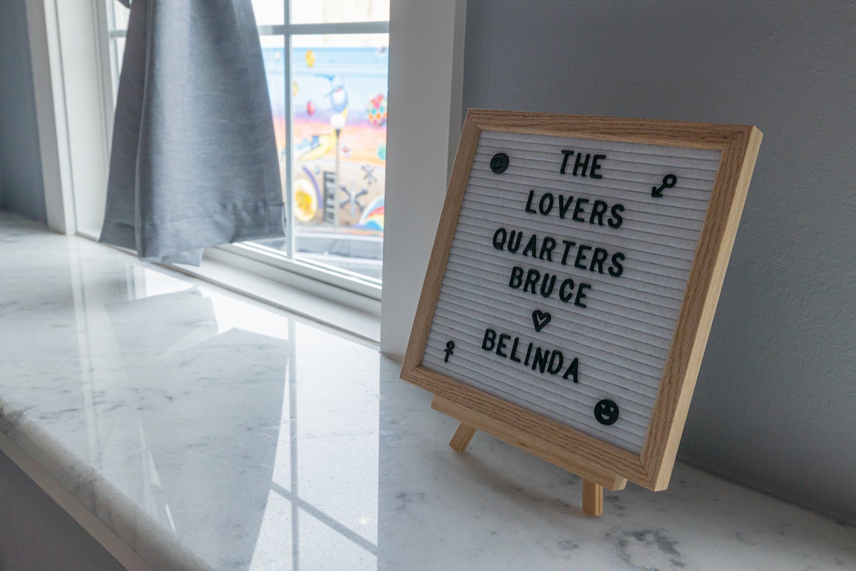 A simple message board placed on a marble countertop displays the names "Bruce" and "Belinda" with decorative symbols. Natural light illuminates the space through a nearby window, which is dressed with a light gray curtain. A colorful mural is visible outside.