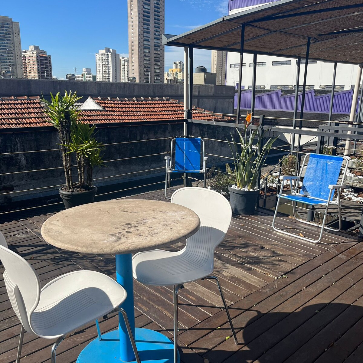 A rooftop deck is highlighted with a round stone table surrounded by three white chairs. Potted plants add greenery, while two blue lounge chairs offer additional seating. The backdrop includes city buildings and a clear blue sky.