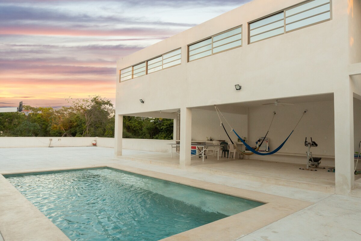 An outdoor pool is featured with clear blue water, surrounded by a spacious concrete area. A cozy hammock is suspended nearby, offering a relaxing spot. The building showcases large windows and a simple white exterior, reflecting the soft evening light against a backdrop of a colorful sky.