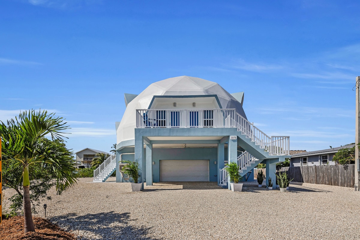 The exterior of a unique spherical house is shown, featuring a light blue façade and a prominent staircase leading to the main entrance. Lush green plants are placed near the base, and a gravel driveway enhances the property’s appeal. Clear blue skies create a serene backdrop.