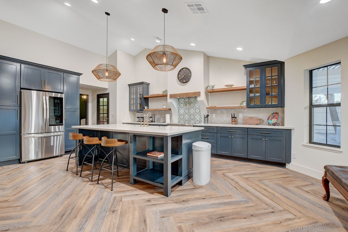 A modern kitchen is highlighted by sleek navy cabinetry and a large island featuring bar stools. Natural light pours in through the windows, illuminating the unique herringbone-patterned wooden flooring. Decorative elements include open shelving and stylish pendant lighting above the island.
