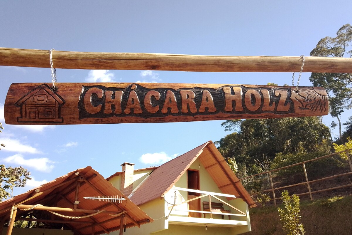 A wooden sign prominently displaying 'CHÁCARA HOLZ' is suspended above a welcoming entrance. The rustic design features carved images of a house and nature. A sloped roof of the nearby building is visible in the background, complemented by a clear blue sky.