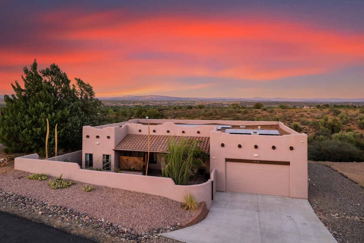 An overview of the home reveals a single-story structure emphasizing traditional Spanish architecture with earthy tones and clean lines. The expansive property is framed by desert landscaping and features a garage, with a sunset sky casting vibrant hues over the scenery.