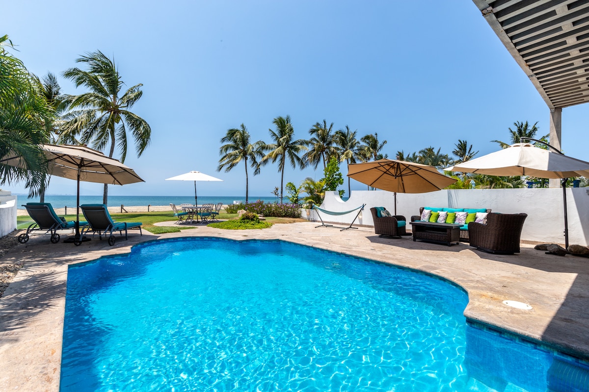 A private swimming pool is surrounded by lounge chairs and umbrellas, with lush palm trees framing the scene. The ocean is visible in the background, just steps away from the pool area, creating a serene outdoor space for relaxation.