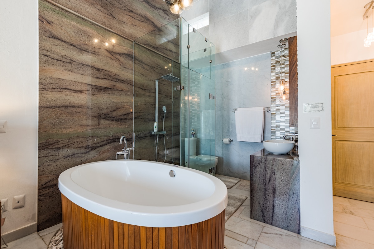 A modern bathroom features a freestanding oval tub with wooden accents. A glass-enclosed shower area is visible, complemented by textured stone walls. Elegant fixtures and a dual-sink vanity enhance the contemporary aesthetic, with natural light illuminating the spacious layout.
