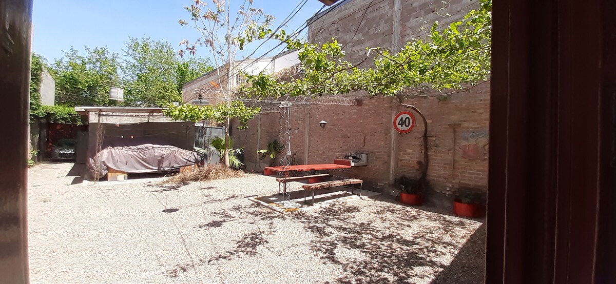 An outdoor patio area is featured, with a large, shaded table set atop gravel. Surrounding plants provide greenery, while a nearby structure offers a covered space. A speed limit sign is displayed, indicating a residential ambiance.