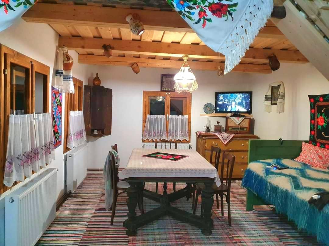 A cozy dining area is featured, highlighted by a wooden table surrounded by chairs. Traditional textiles adorn the walls, while a vintage cabinet and a television are visible. Natural light streams through the windows, enhancing the warm ambiance of the space.