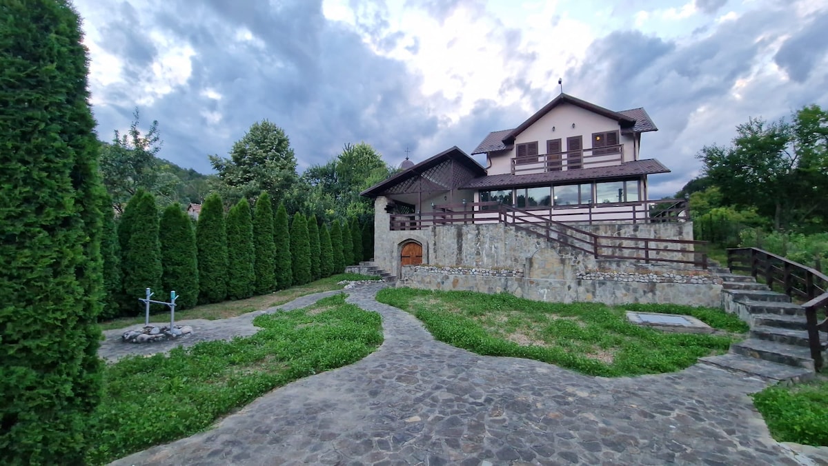 A two-story house with a wooden façade is surrounded by neatly trimmed hedges and a stone pathway. Lush greenery leads to a spacious lawn, while a wooden deck provides outdoor seating options. Cloudy skies create a serene backdrop for this nature-centered retreat.