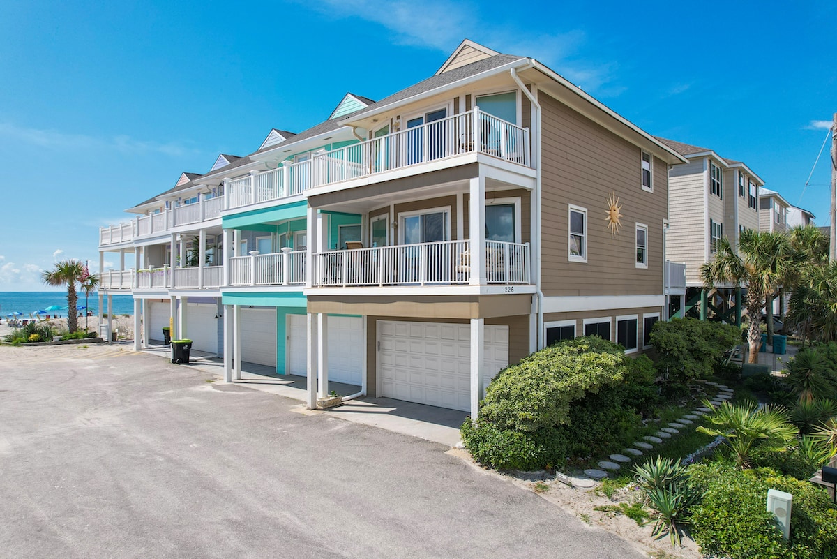 A multi-level beach house is shown, featuring a light-colored exterior with balconies and a sun emblem. The property is positioned near the ocean, surrounded by palm trees and greenery. An enclosed two-car garage is visible, along with a spacious driveway leading to the entrance.