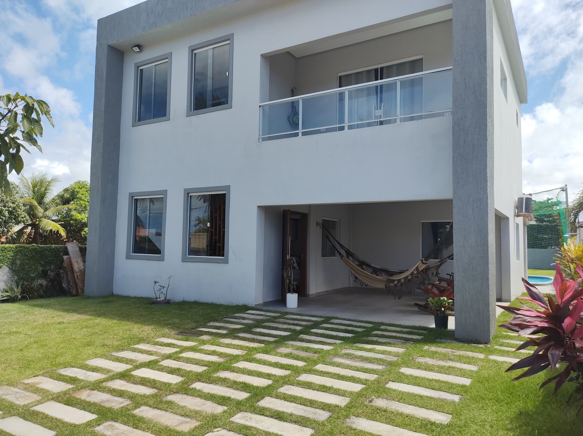 A modern house stands surrounded by a well-maintained lawn. The entrance features a hammock hanging on the porch, while large windows provide natural light. A stone path leads to the front door, enhancing the ease of access to the outdoor spaces.