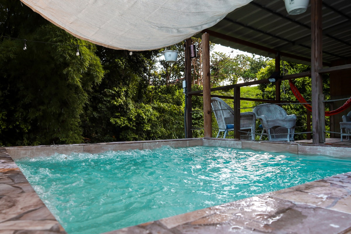 A private pool area is visible, surrounded by natural greenery. The water features a bubbling feature and is partially covered by a light tarp. Two wicker chairs are positioned nearby, creating a cozy setting for relaxation.