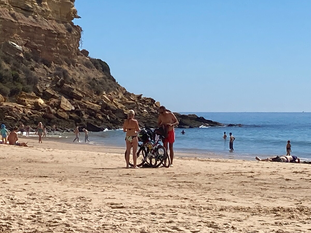 A sandy beach is depicted with people enjoying the sun and water. Two individuals stand near a bicycle, surrounded by others relaxing or swimming. The backdrop features a rocky cliff, while the calm sea stretches into the distance under a clear blue sky.