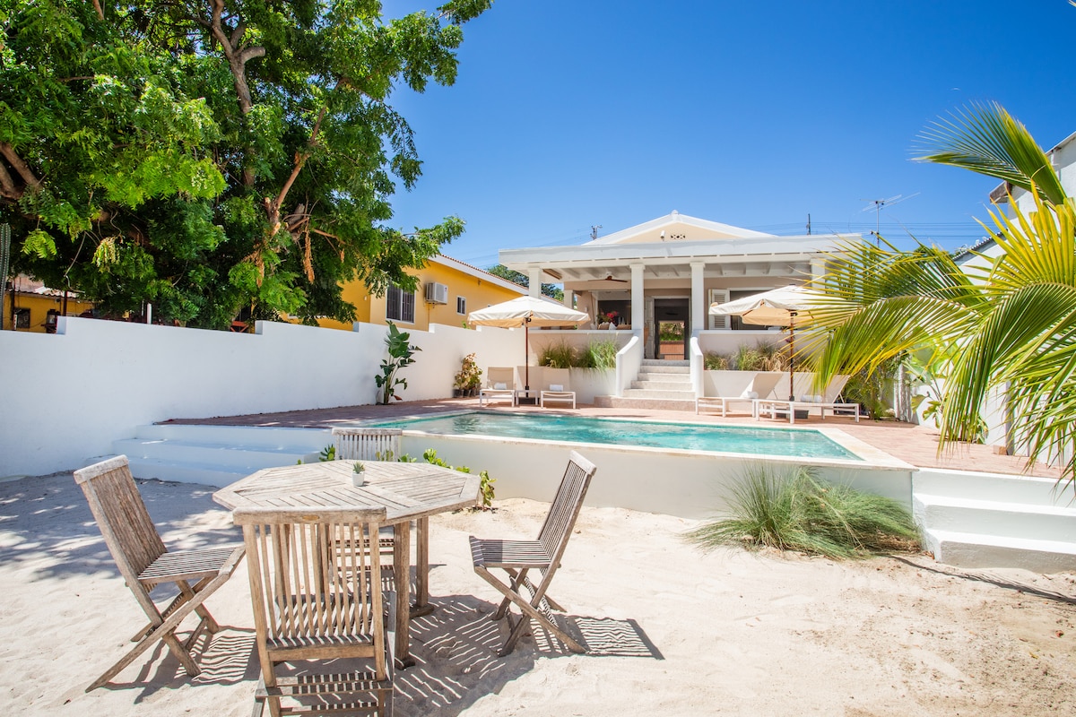 A spacious outdoor area features a wooden dining table with accompanying chairs, surrounded by sand and greenery. A private mineral-based pool is visible in the background, with comfortable lounge chairs placed under sun umbrellas, offering a relaxing space near the beach house.