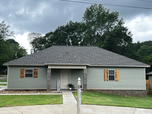 A single-story home with a light green exterior features a central entrance framed by two decorative shutters. The roof is dark gray, and the front yard is landscaped with grass and small rocks. A smooth driveway leads up to the entrance.