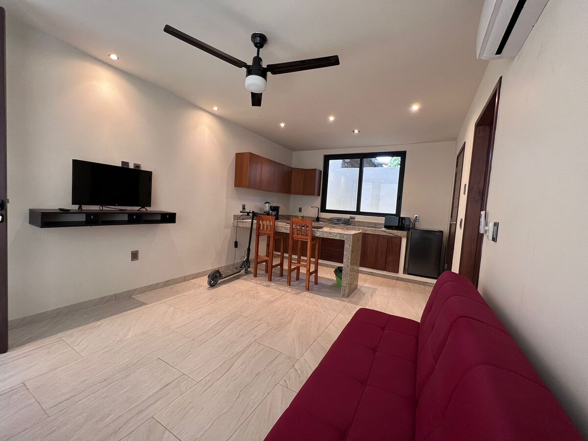 A well-lit living area features a comfortable red sofa paired with two wooden chairs at a counter. A TV is mounted on the wall, while a modern kitchen is visible in the background, equipped with essential appliances. An electric scooter is parked nearby.