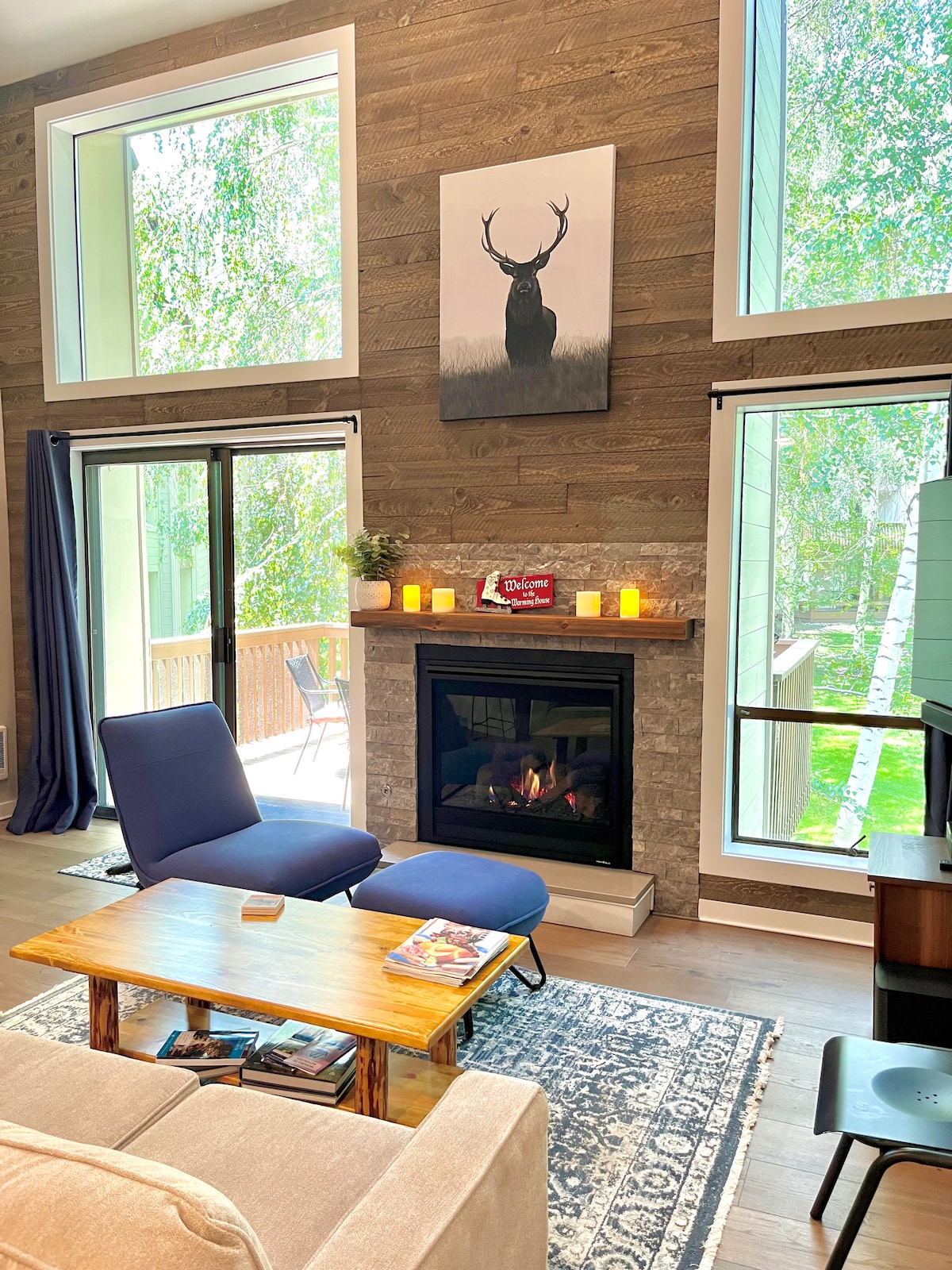 A welcoming living space features large windows that invite natural light. A comfortable seating arrangement includes two blue chairs and a wooden coffee table, positioned near a stone fireplace. A nature-themed artwork adorns the wall above, while soft lighting creates a calming ambiance.