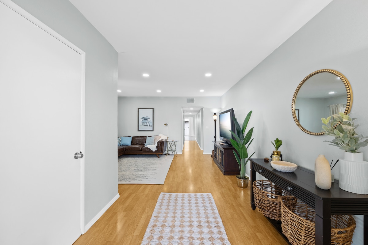 A welcoming living area features smooth wooden flooring and soft grey walls. A decorative mirror and potted plant adorn a side table, while a television is set against an elegant media console. Light from recessed lighting enhances the space, which opens to additional rooms.