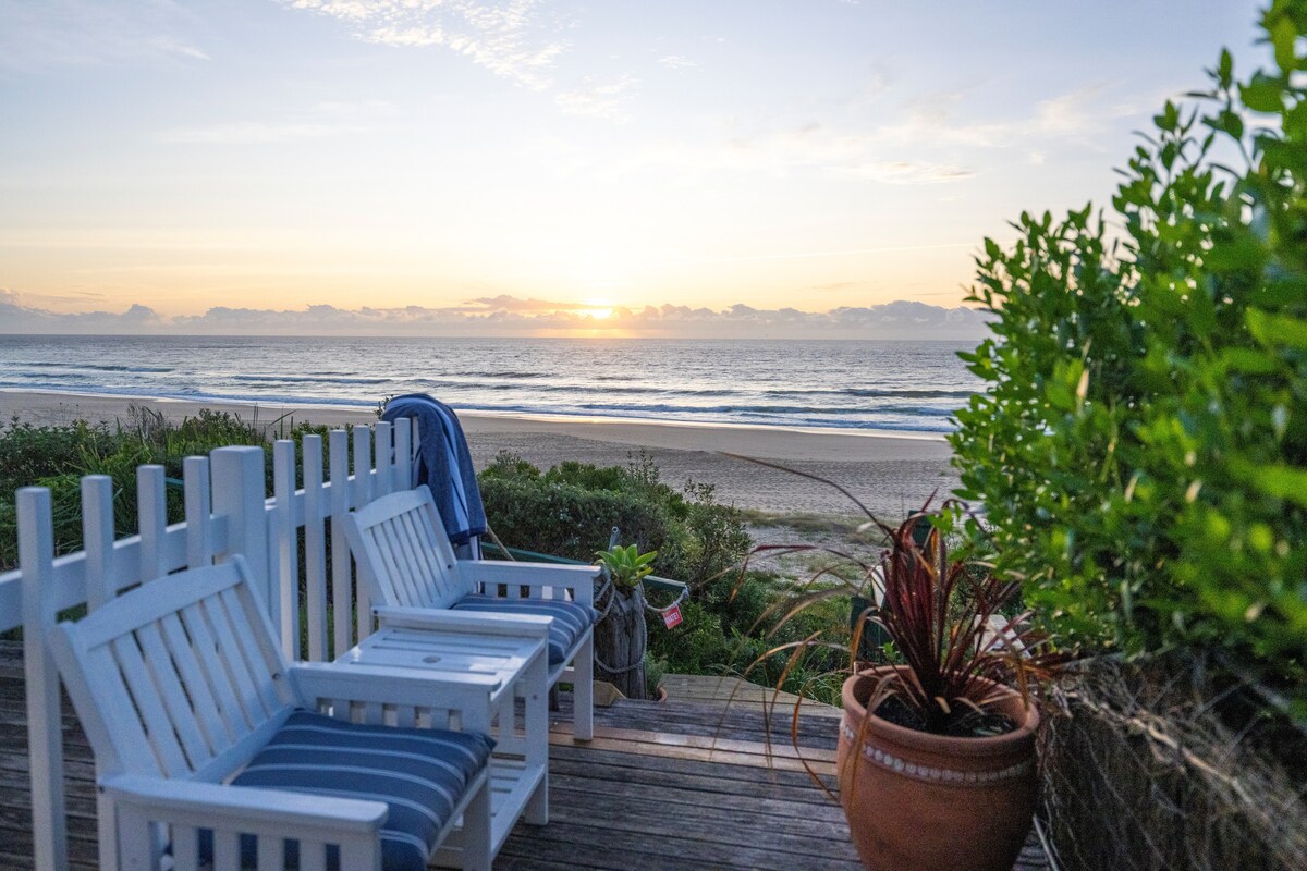 A beach deck is presented with two white chairs facing the ocean. The sun begins to rise above the water, illuminating the sky with soft colors. Lush greenery enhances the view, providing a peaceful transition from the deck to the beach.