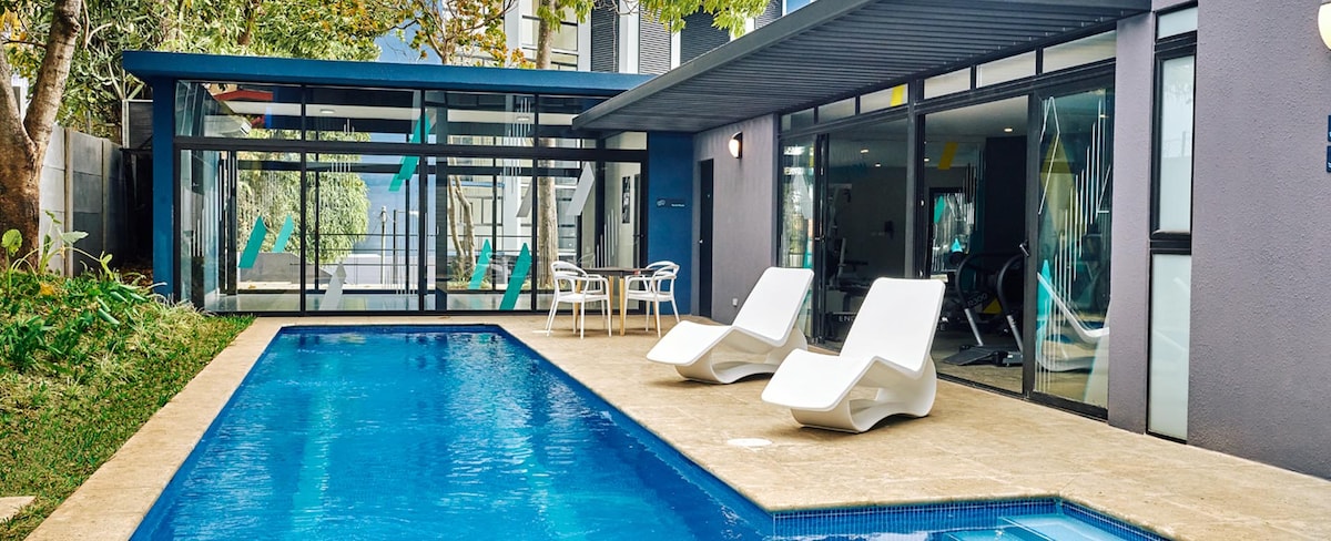 An inviting outdoor space features a swimming pool bordered by a light-colored patio. Two modern lounge chairs are positioned beside the water, and large sliding glass doors provide views into the living area. Lush greenery enhances the serene atmosphere.