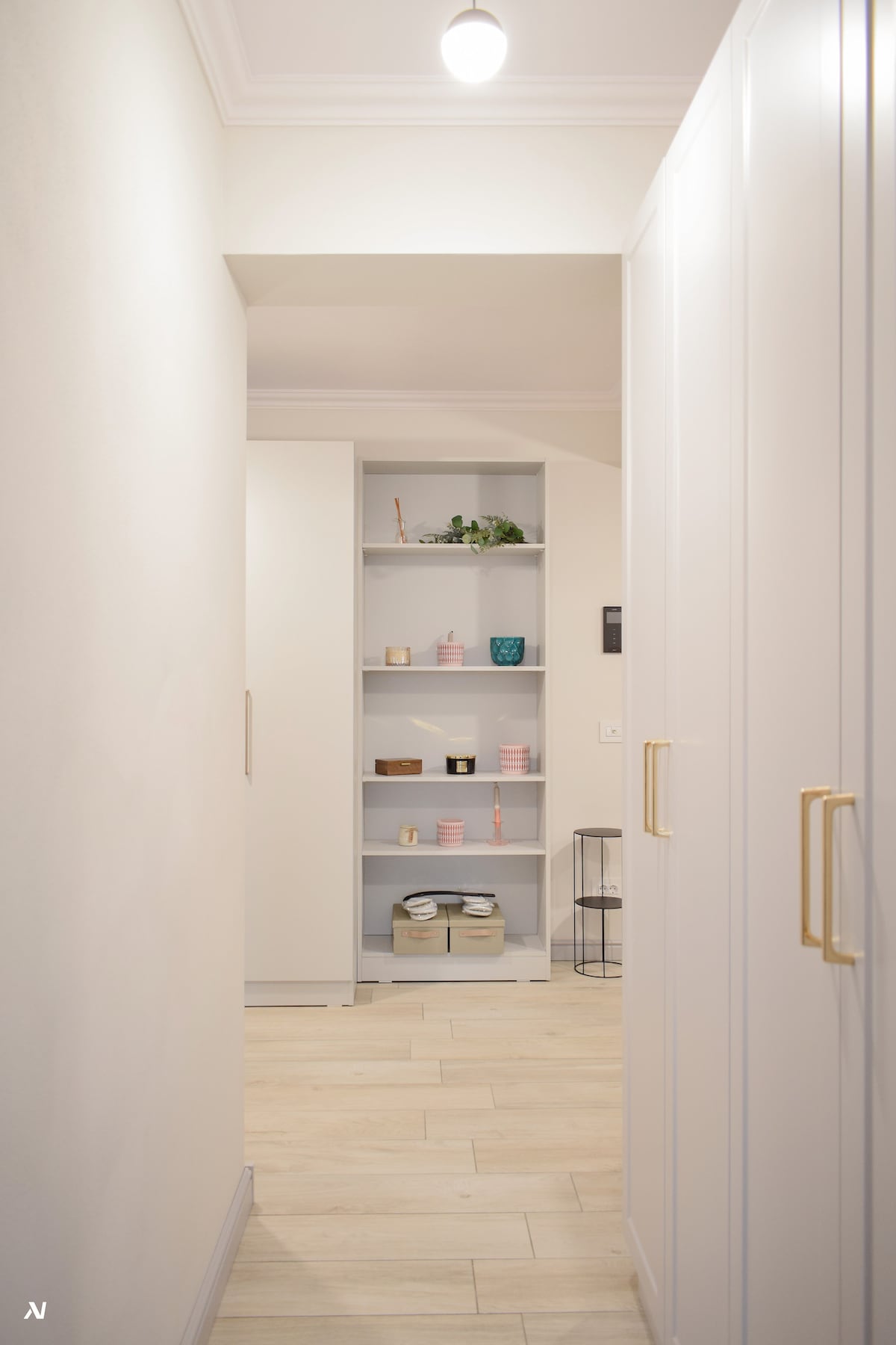 A well-organized hallway is featured, with sleek white cabinetry lining the sides. A small shelf is visible in the background, showcasing neatly arranged decorative items and storage bins. Soft natural light illuminates the space, creating a calm and tidy atmosphere.
