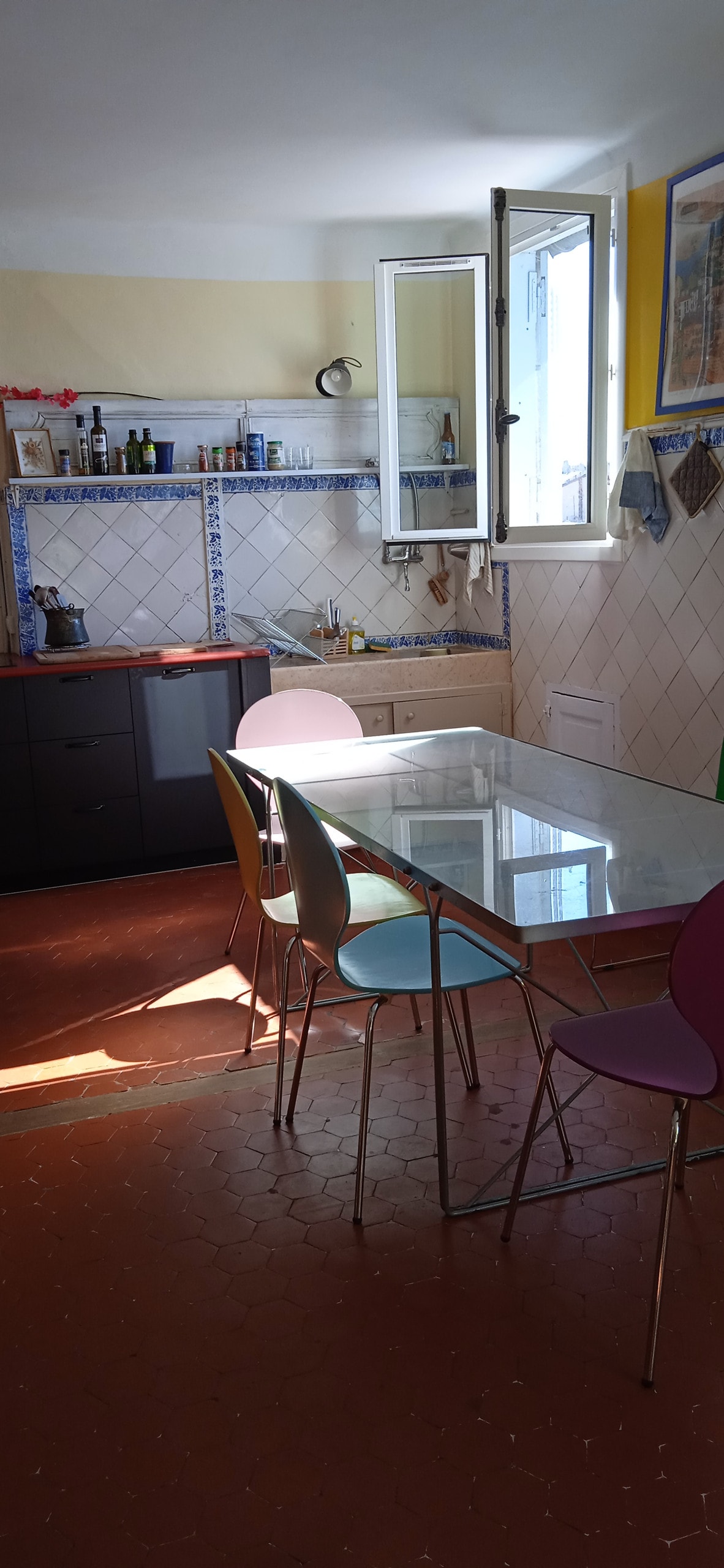 A kitchen space is illuminated by natural light from a window, showcasing a glass dining table surrounded by colorful chairs. Cabinets and a countertop are visible, with kitchenware neatly arranged on the shelves above. The floor features warm, patterned tiles.
