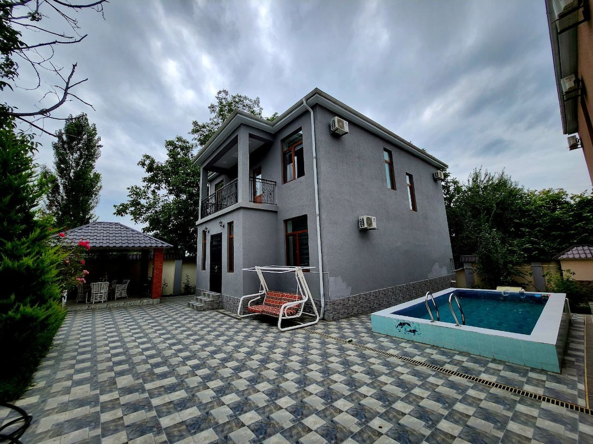 A two-story villa is shown with a modern exterior, featuring large windows and a balcony. An outdoor swimming pool is visible beside the villa, surrounded by a tiled patio. Lush greenery is present around the property, contributing to a tranquil setting.