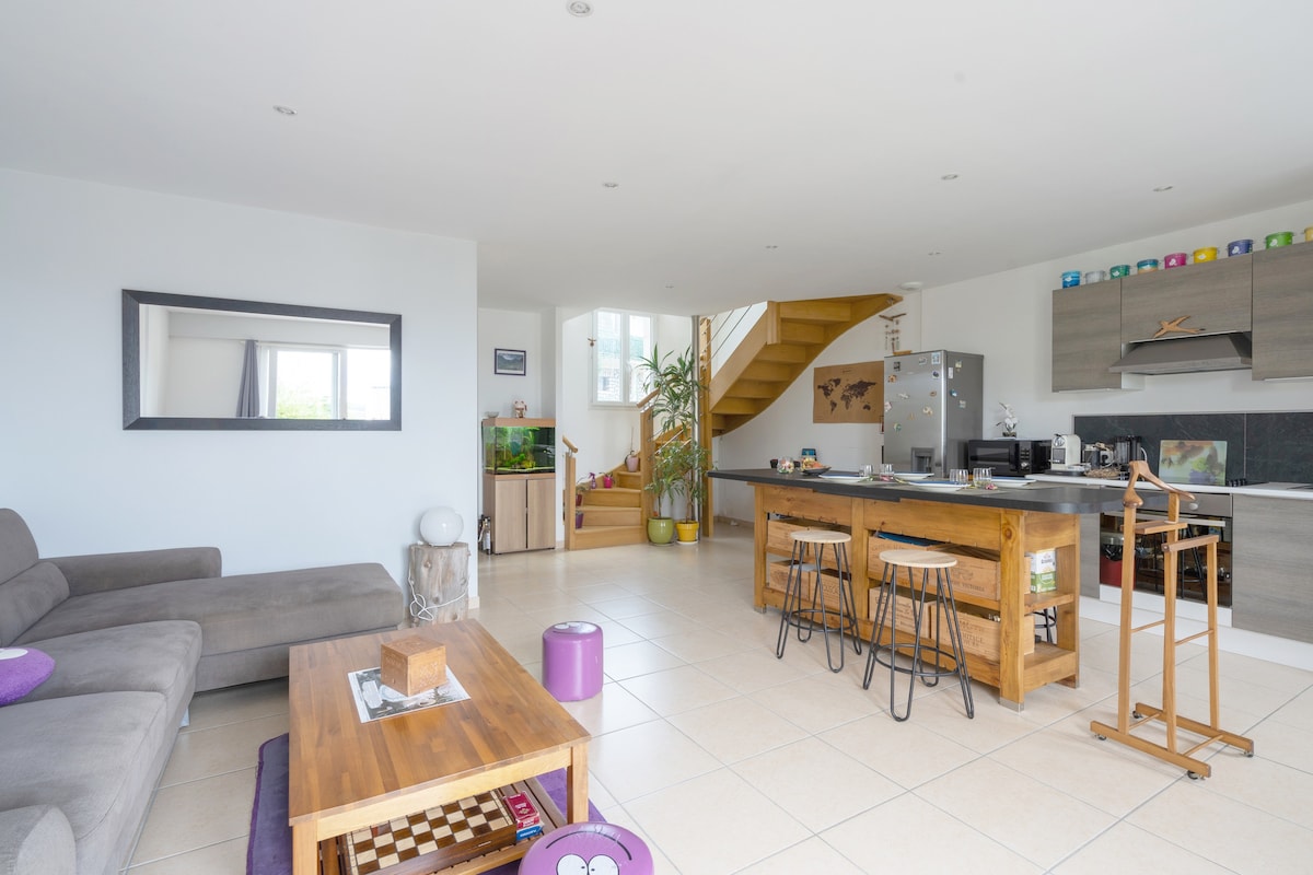 The spacious living area features a comfortable gray sofa and a wooden coffee table, with light tiles enhancing the open layout. A modern kitchen is visible, equipped with appliances and bar stools. Large windows allow natural light to fill the space, complementing the stylish decor.