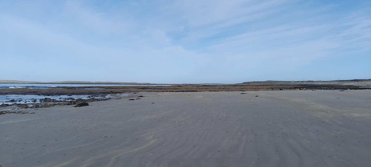 A wide sandy beach stretches into the distance, with gentle waves lapping at the shore. The calm blue sky reflects on the water, while distant rocky outcrops are visible. The beach offers ample space for walking and exploration, creating a serene coastal atmosphere.
