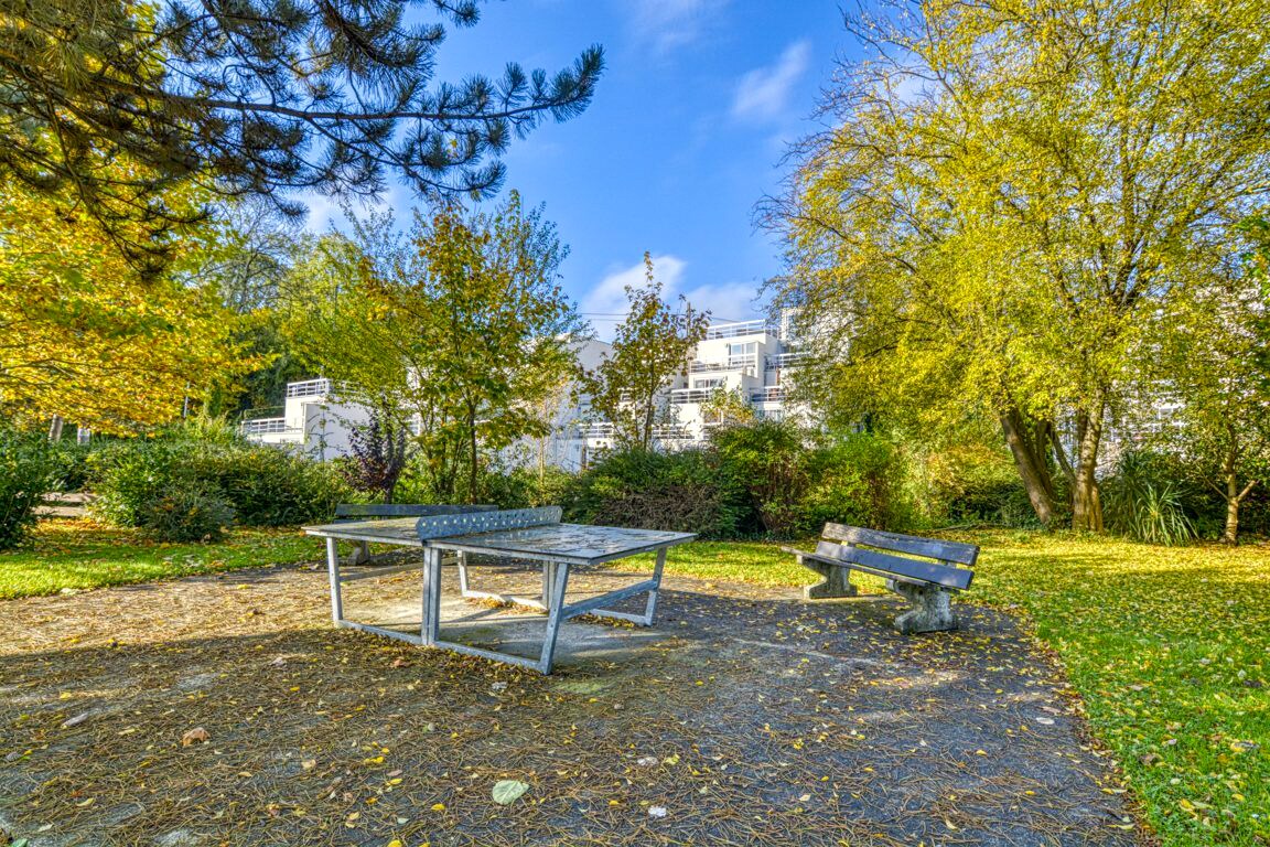 A park area features a table suitable for outdoor activities and two benches surrounded by greenery. Trees with yellowing leaves provide shade, while a few scattered leaves cover the ground. Light filters gently through the foliage, creating a serene environment.