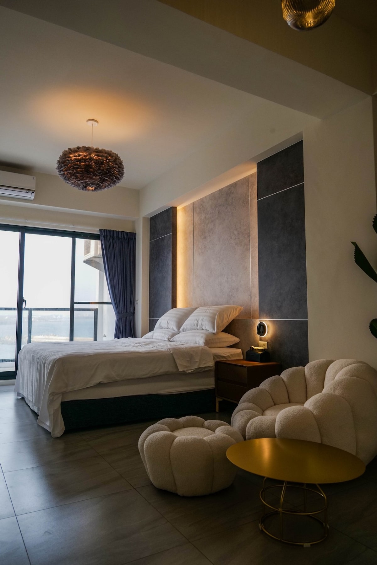 A well-appointed bedroom features a spacious bed positioned against a textured wall, with warm lighting accentuating the decor. A cozy armchair and a small gold table provide comfortable seating. The large window offers scenic views, allowing natural light to brighten the space.