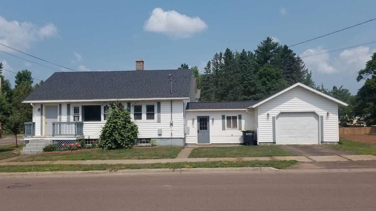 The exterior of a newly remodeled 1930's home is displayed, featuring a white façade, a dark roof, and a welcoming porch. A manicured lawn enhances the curb appeal, while a garage is positioned on the right. Lush trees fill the background, providing a sense of privacy.