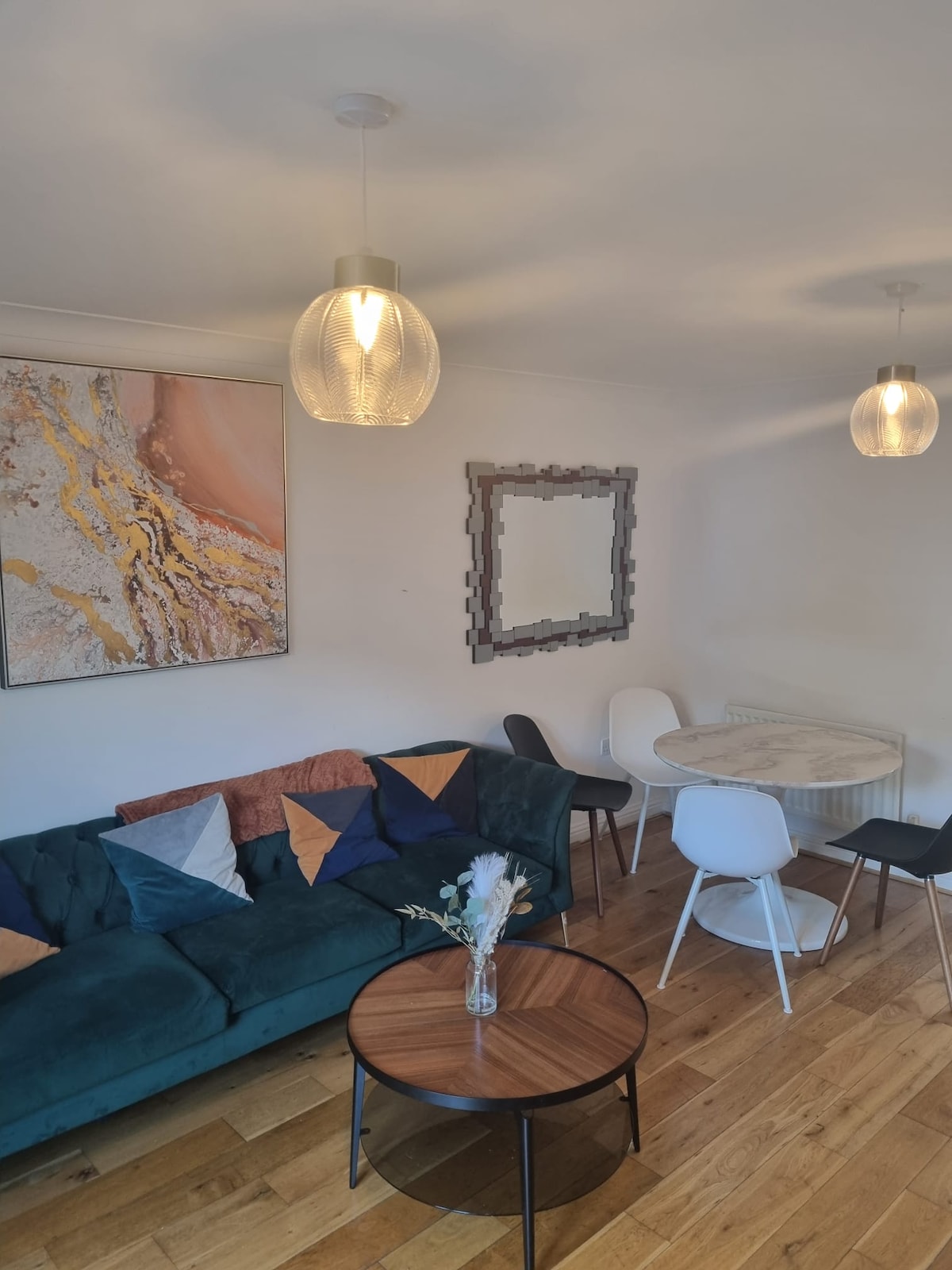 A modern living space features a green sofa adorned with various colored cushions. A round dining table with white and black chairs is situated beside the wall. A large abstract artwork hangs above the sofa, and two pendant lights illuminate the room.