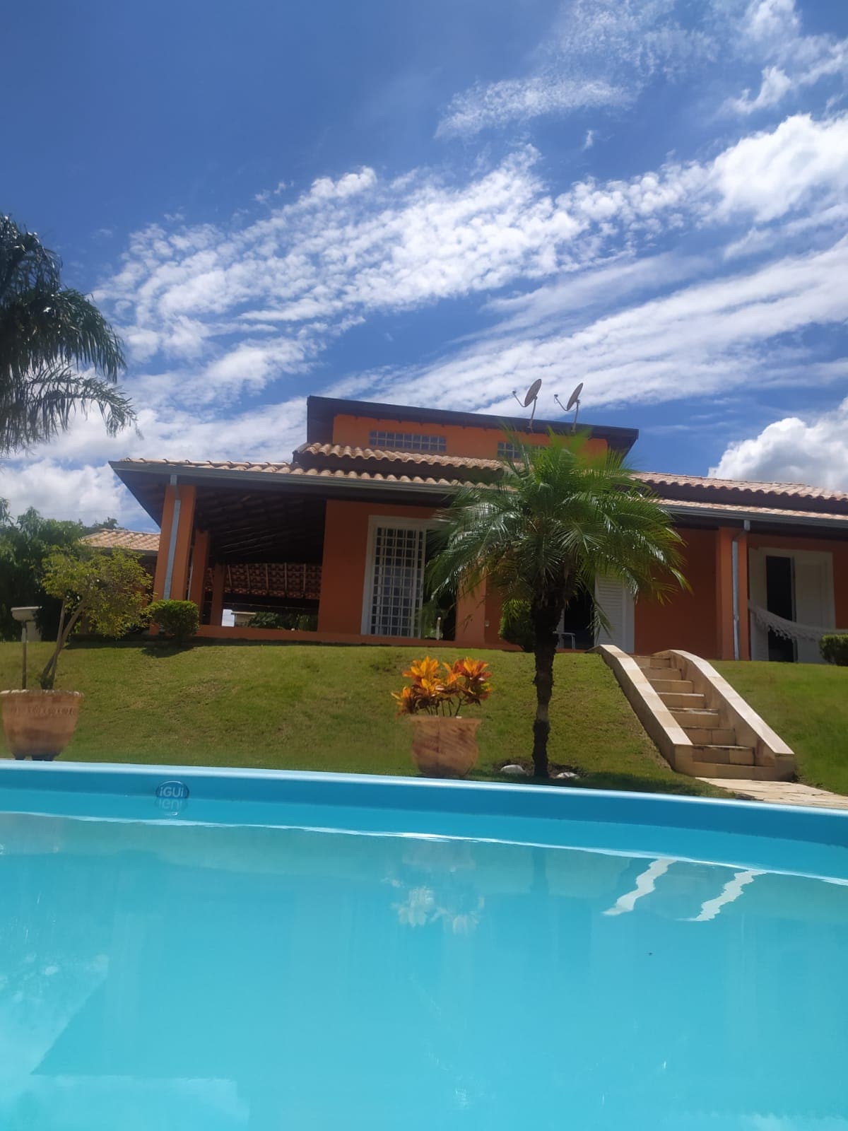 The image captures a vibrant orange building with a terracotta roof, set against a bright blue sky. A swimming pool reflects the structure, while lush greenery and potted plants adorn the surrounding area, enhancing the serene outdoor environment.