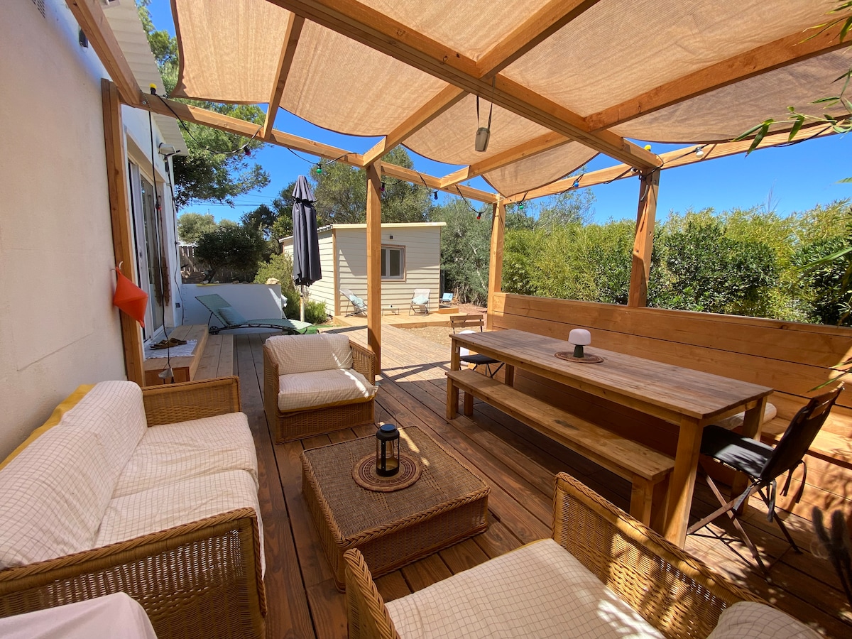 An outdoor terrace is surrounded by a wooden structure with a canopy providing shade. A large dining table and several seating areas are visible, featuring natural wood textures. Lush greenery can be seen in the background, contributing to a serene environment.