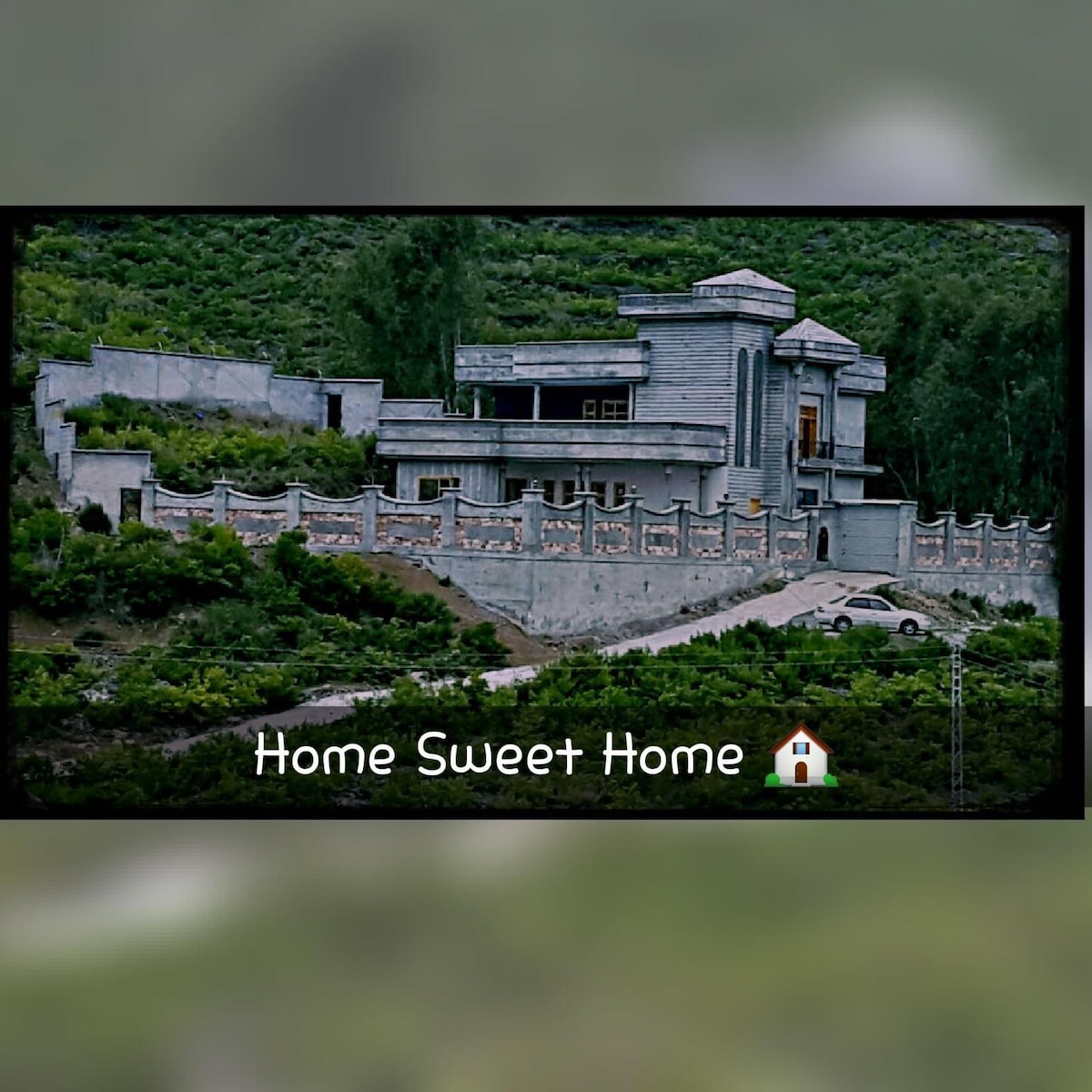 A contemporary two-story home is set against a lush green hillside. Light-colored stone walls and a gabled roof create a modern aesthetic. A driveway leads to the entrance, where two cars are parked. A decorative wall surrounds the property, enhancing its privacy.