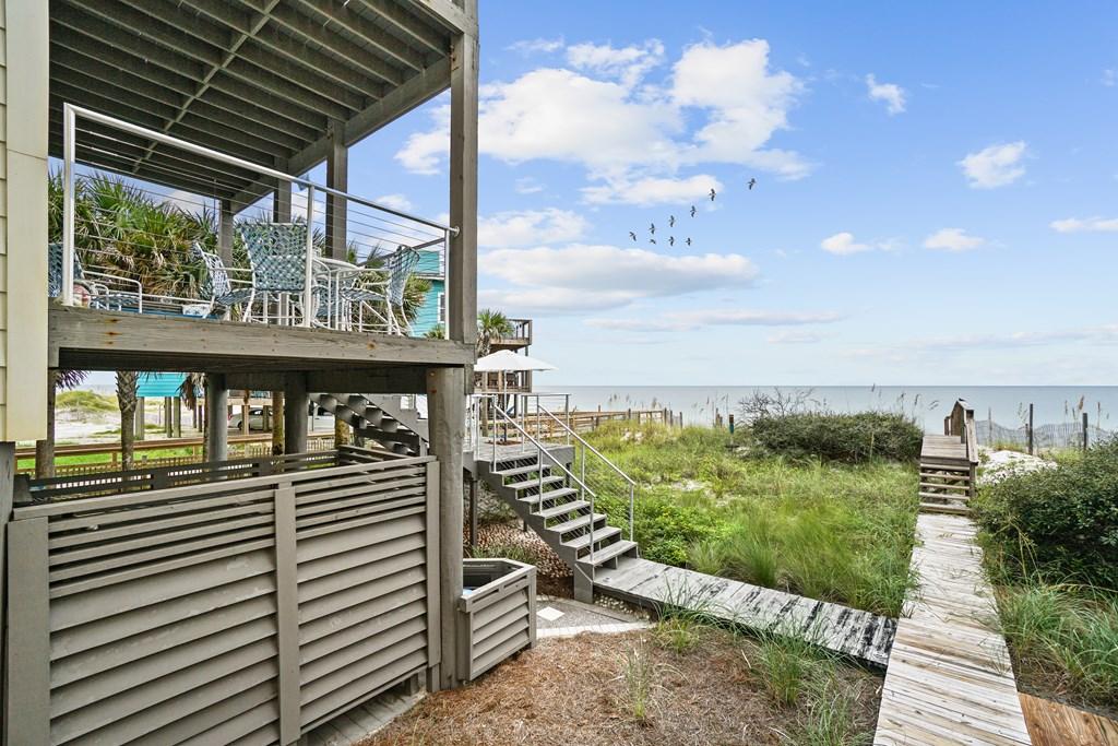 Two decks are visible from this outdoor view, one featuring a dining set. A wooden pathway leads to the beach, framed by tall grass and sandy areas. The ocean horizon is seen in the distance under a clear blue sky, complemented by scattered clouds.