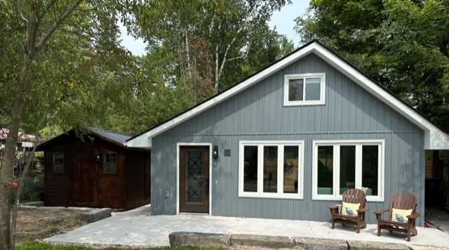 A charming exterior of the home features a gray facade with large windows that invite natural light. Two wooden Adirondack chairs are placed on a concrete walkway, framed by lush greenery and trees. A wooden shed is situated to the left of the house.