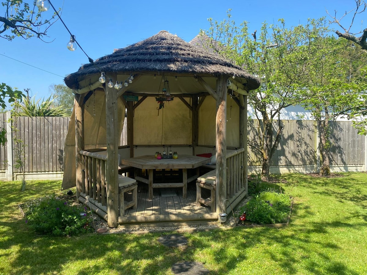 A round thatched gazebo is set in a lush green garden, surrounded by well-maintained grass and a wooden fence. The gazebo features a wooden table surrounded by benches, with decorative plants and flowers positioned around the perimeter. Soft lighting hangs overhead, creating a serene atmosphere.