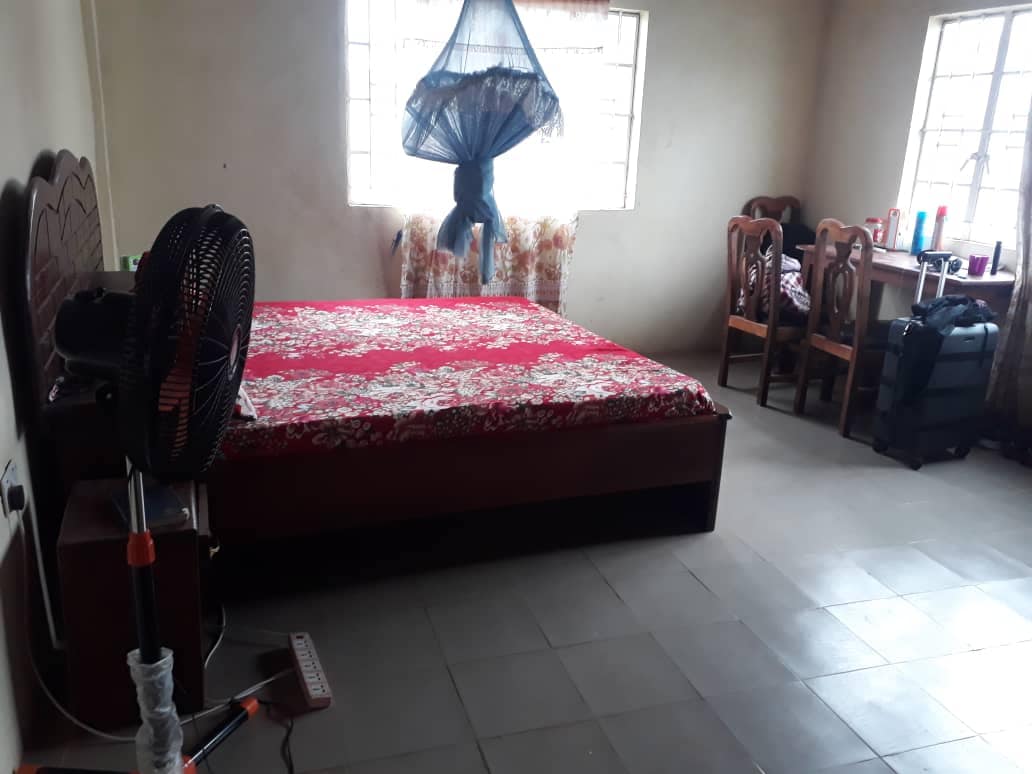 A spacious room features a red floral-patterned bedspread on a large bed. Two windows provide natural light, with sheer curtains and a cotton canopy above the bed. A wooden table with chairs stands in one corner, and a standing fan is positioned nearby.