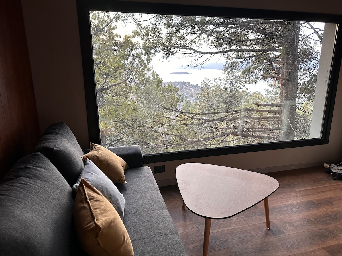A modern seating area is highlighted by a comfortable sofa adorned with cushions. A minimalist wooden coffee table sits in front, while a large window provides a scenic view of the surrounding forest and lake. Natural light fills the space, creating an inviting atmosphere.