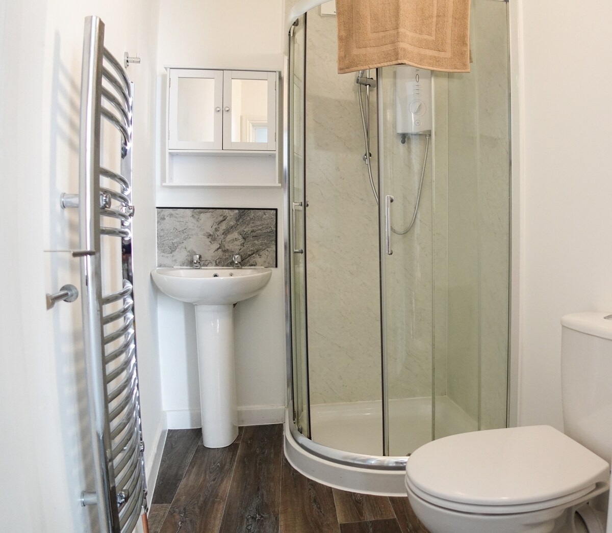 A clean and modern ensuite bathroom features a walk-in shower with transparent glass doors. A white sink is positioned next to the shower, with a large mirror above. The towel rack and the toilet are also visible, set against a neutral wall and wooden floor.