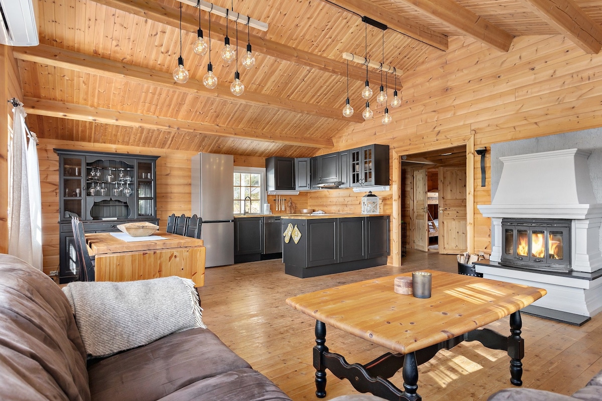 A spacious living area features wooden walls and high ceilings with hanging lights. A large dining table is paired with black chairs, while a modern kitchen with dark cabinetry is visible in the background. A cozy fireplace adds warmth to the room, enhancing the inviting ambiance.