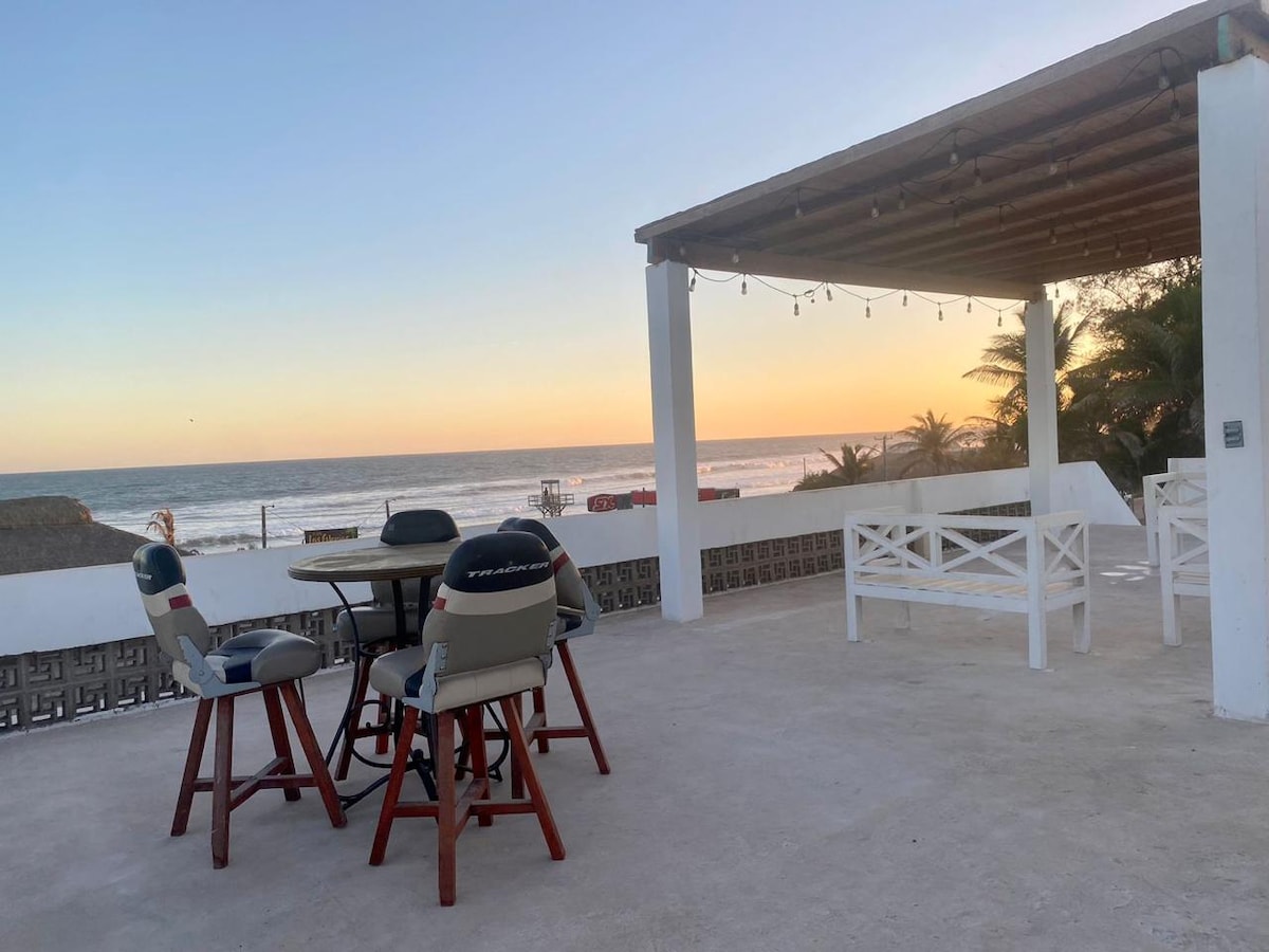 A spacious outdoor terrace features a circular wooden table surrounded by high-backed chairs, providing a place to relax. A sunset view over the ocean is visible, with soft colors filling the sky and palm trees lining the background, enhancing the coastal experience.
