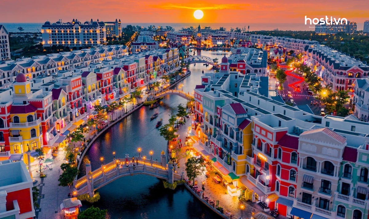 A vibrant view of a waterfront neighborhood is captured during sunset, with colorful buildings lining the canals. The scene features a pedestrian bridge illuminated by warm lights, reflecting on the water, while the setting sun casts a golden hue over the landscape.