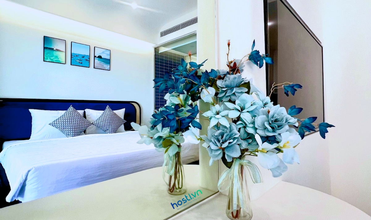 A spacious sleeping area features a neatly made bed with subtle blue and white accents. Decorative floral arrangements are placed in vases on a vanity, complementing the modern design. A large television and elegant wall art add to the inviting atmosphere.