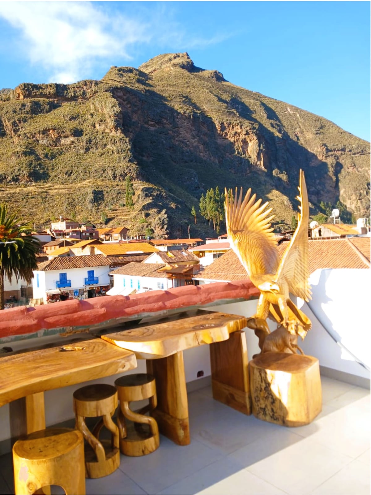 A wooden table with matching stools is positioned on a terrace, offering views of a mountainous landscape. A large carved eagle sculpture is prominently featured beside the table. The backdrop includes terracotta-roofed buildings nestled in the valley below.
