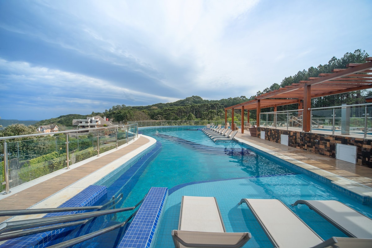 An outdoor pool area is displayed, featuring clear blue water and several lounge chairs positioned along the edge. A wooden pergola provides shaded seating, while lush green hills are visible in the background under a cloudy sky.